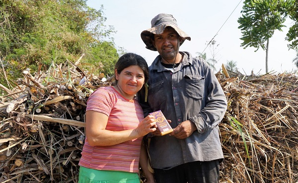 Two sugar co-op members hold up a chocolate bar in front of stacks of sugarcane.