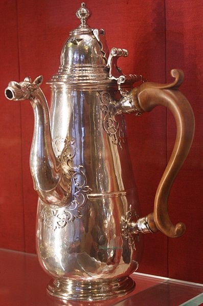 Elaborately molded silver pot has a spout, attached lid and a handle of a metal that looks like brass.