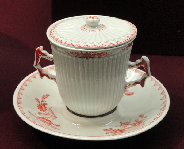 White china cup resembles a tea cup, but has two handles and a lid as well as a saucer, all with a red floral pattern.