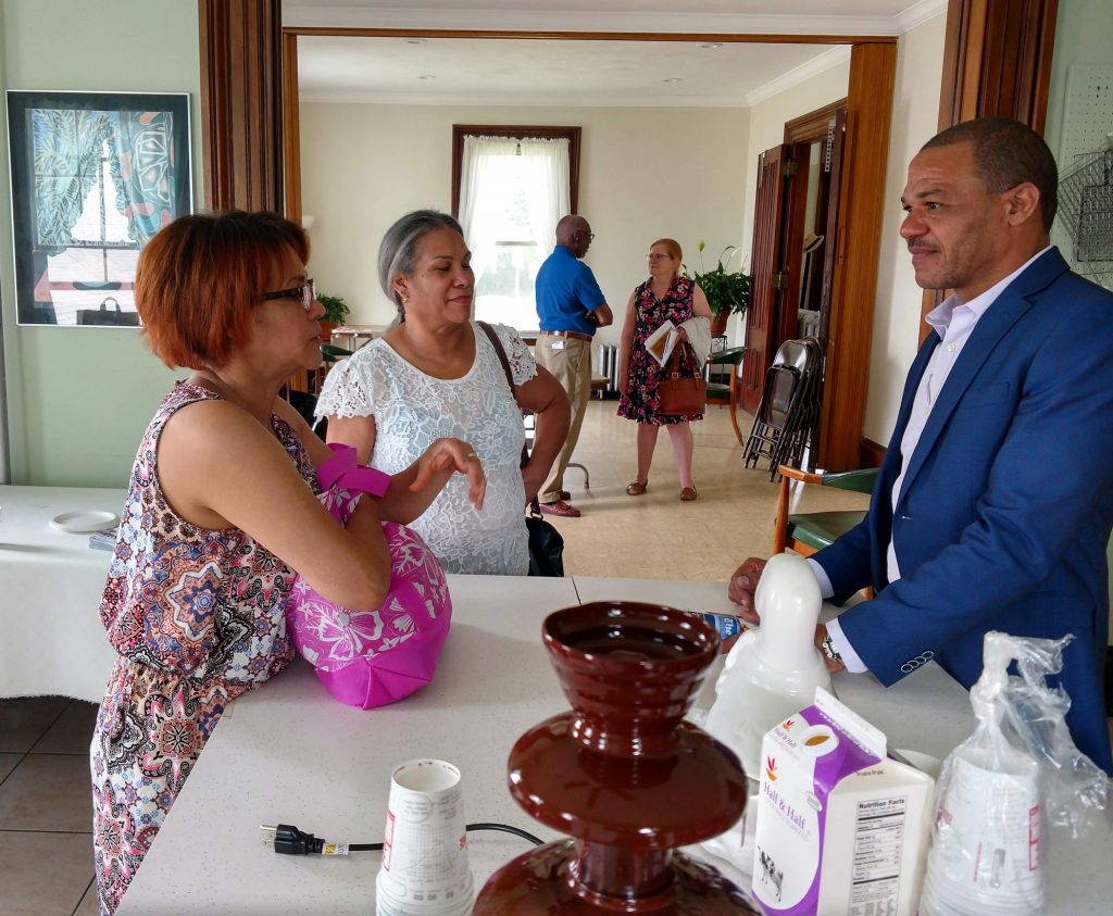 A man, Abel Fernandez, talks to two women about fair trade cacao. A chocolate fountain sits in the foreground.
