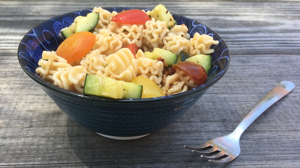 a bowl of pasta salad with vegelables sits on a wooden table next to a fork