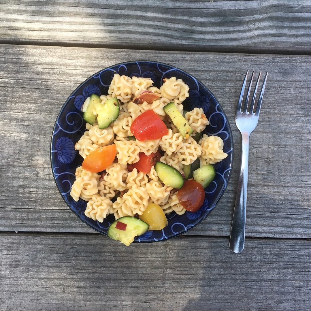 a bowl of pasta salad with vegelables sits on a wooden table next to a fork