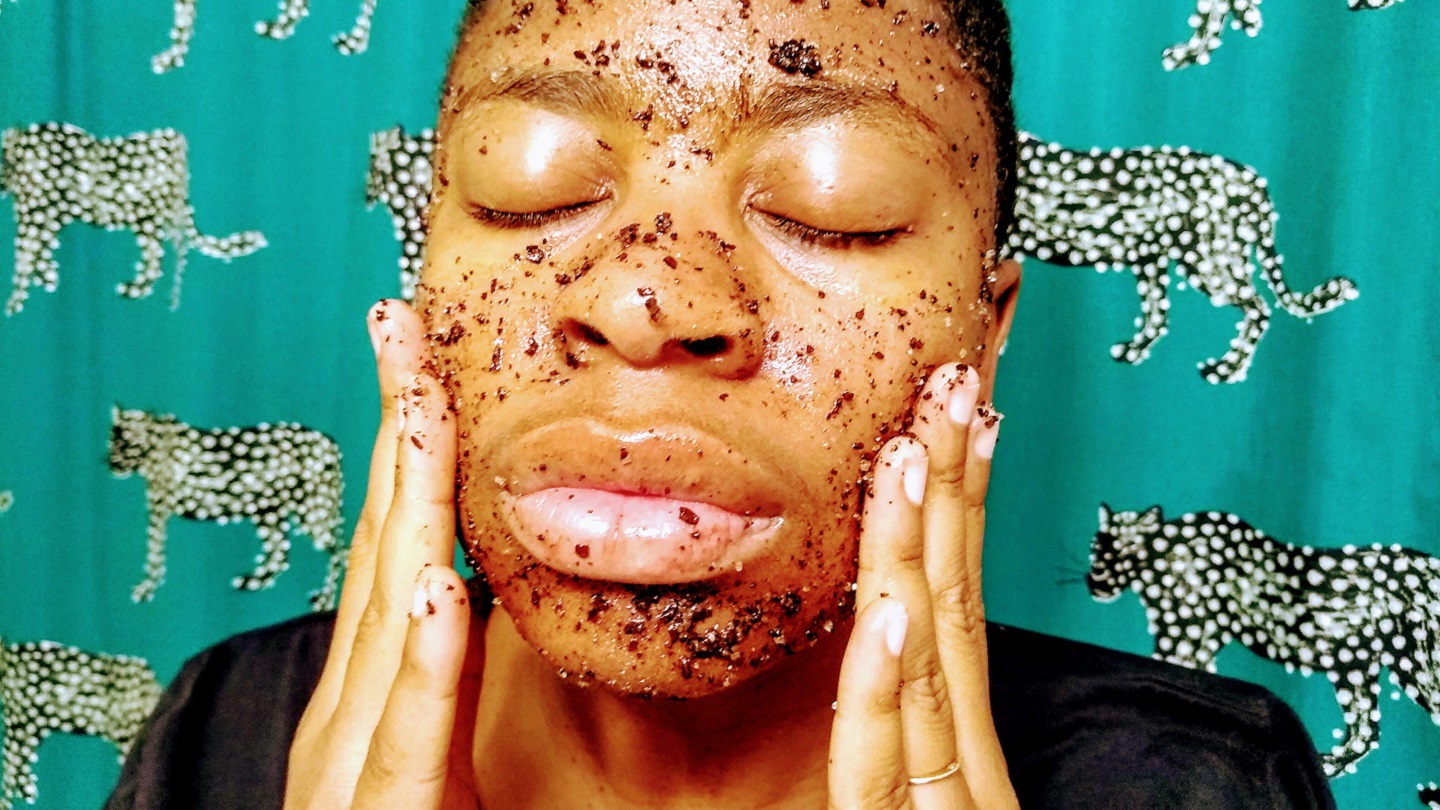 A woman with a coffee scrub on her face rubs her cheeks with her fingertips