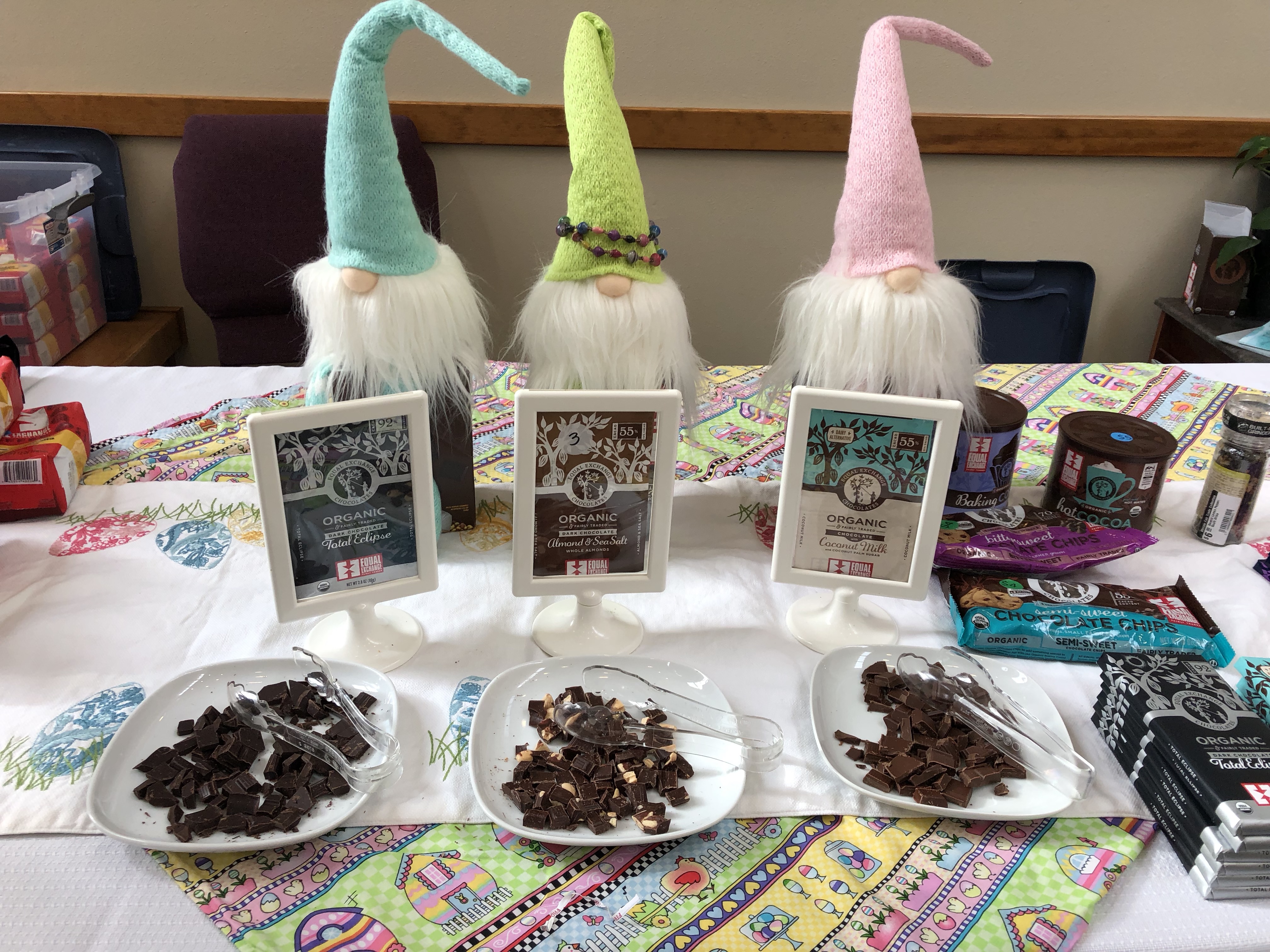 Three gnome dolls sit in front of three plates of chocolate piece samples on plates with tongs.