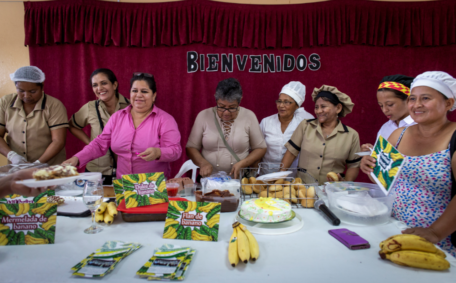 Group of women cooks at a table full of bananas and cakes, under a sign reading 'Bienvenidos'